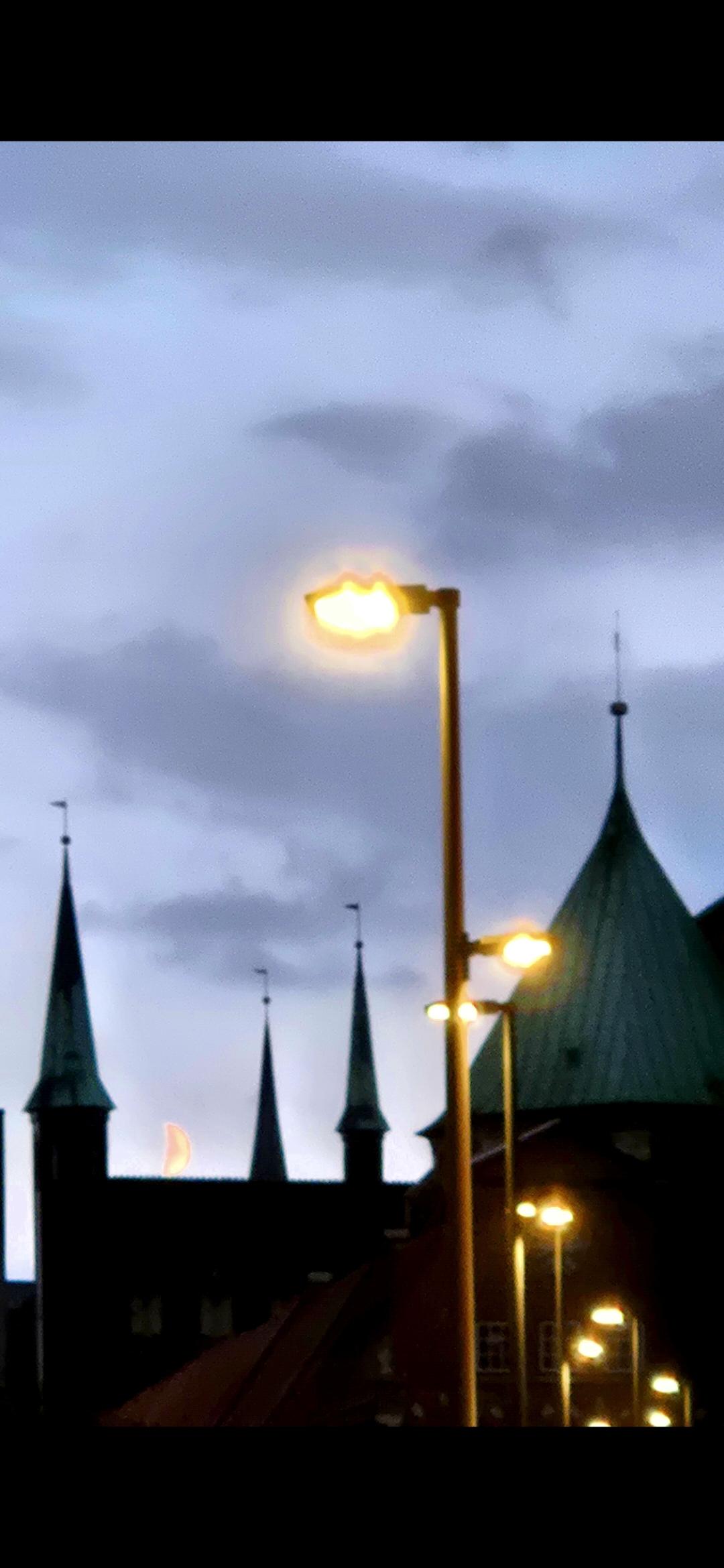 Mondsichel hinter Kirchtürmen, im Vordergrund Straßenbeleuchtung.
