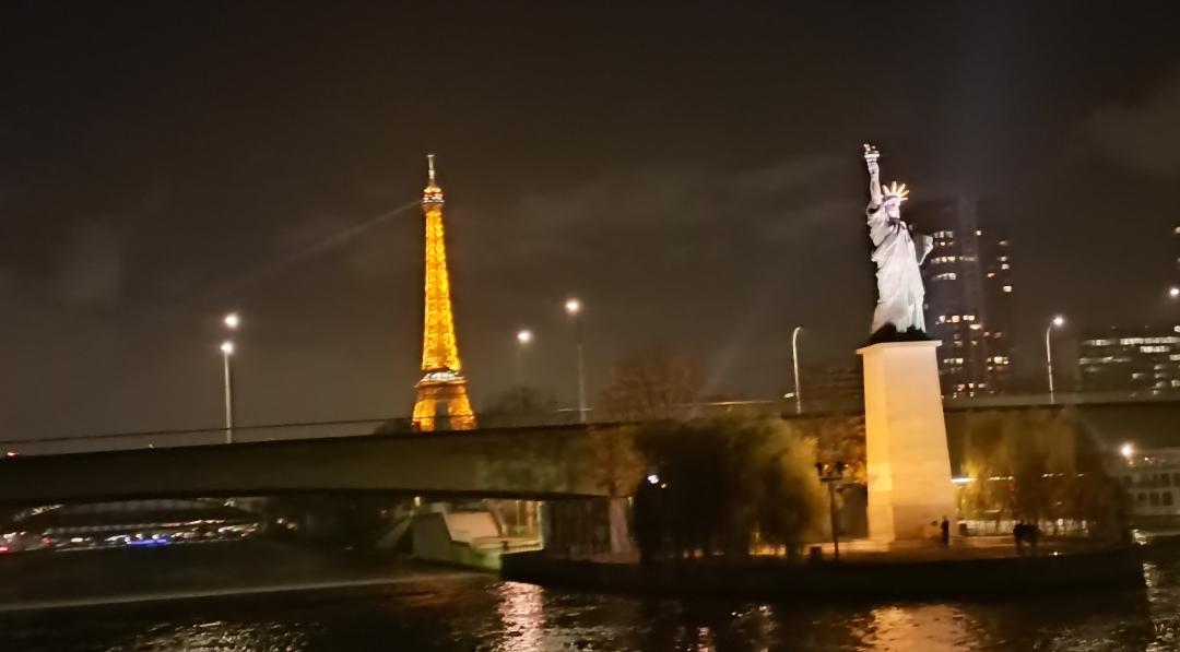 Photo très floue et de traviole, car de nuit et prise de loin, de la Seine à Paris, montrant la Tour Eiffel illuminée et la reproduction de la statue de la liberté éclairée. 