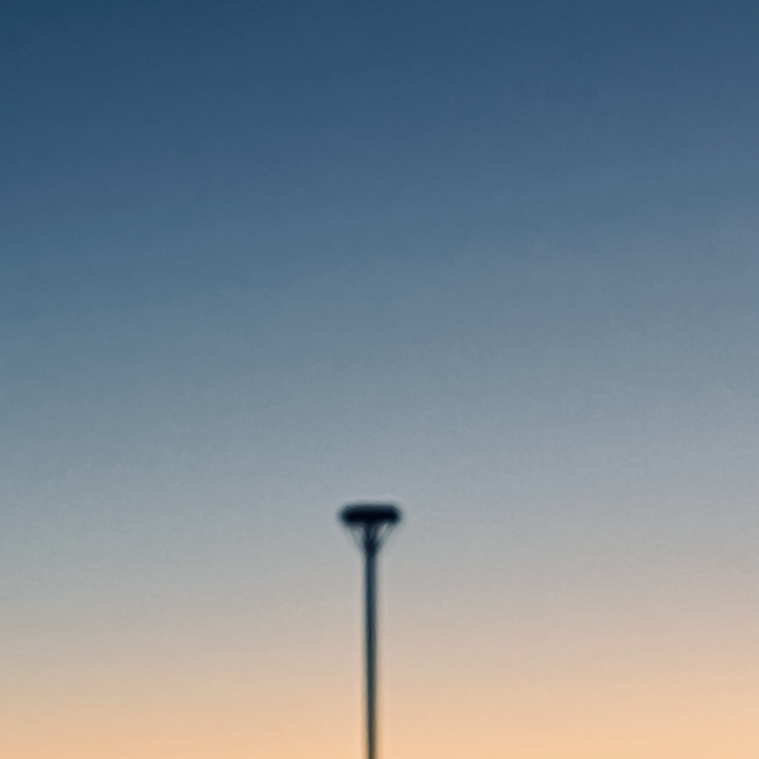 Im Abendrot verläuft die Himmelsfärbung von Hellorange nach Stahlblau. Senkrecht ragt ein Pfahl schwarz in das Bild auf dem oben ein Plateau montiert ist 