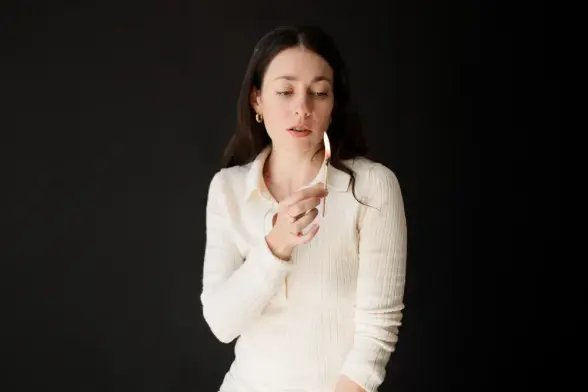 Lia Kohl in a white sweater holds a lit match, set against a dark background, creating a focused and contemplative mood.