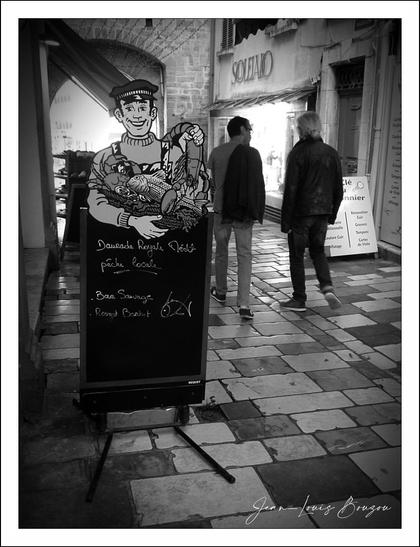 The image captures a charming street scene in black and white, evoking a timeless and somewhat nostalgic atmosphere. At the forefront, there is a prominent sandwich board featuring an illustration of a sailor or fisherman, dressed in a classic striped shirt and a cap. He holds a large lobster or crab, symbolizing fresh seafood, and his cheerful expression invites passersby to explore the delicacies offered.
Written on the board in elegant, cursive handwriting are the daily catches available.
In the background, two people walk away from the camera along a cobbled street, adding a sense of everyday life and movement to the scene. The texture of the stone pavement and the surrounding rustic architecture add to the old-world charm and likely indicate a historic or coastal town, known for its seafood tradition.
The image’s monochrome tone enhances the mood, focusing attention on contrasts of light and shadow and allowing the details of the drawing and the street to stand out. Symbolically, the sailor with fresh seafood can represent themes of tradition, hard work, and the bounty of the sea, appealing to both locals and tourists alike. Overall, this photograph artfully combines human presence, artistic signage, and the essence of place to evoke a story of culinary culture rooted in maritime heritage.
