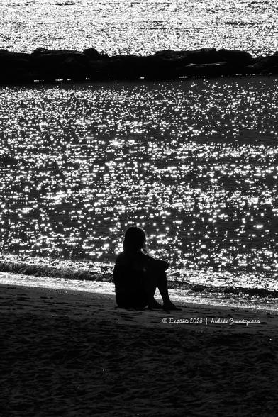 Imagen vertical de una mujer sentada frente al mar sola. Aparece en el tercio inferior y centrada de la fotografía. Debajo está la arena y, en los tercios superiores, el mar con muchos destellos y un trozo de espigón que atraviesa el cuadro de un lado a otro.