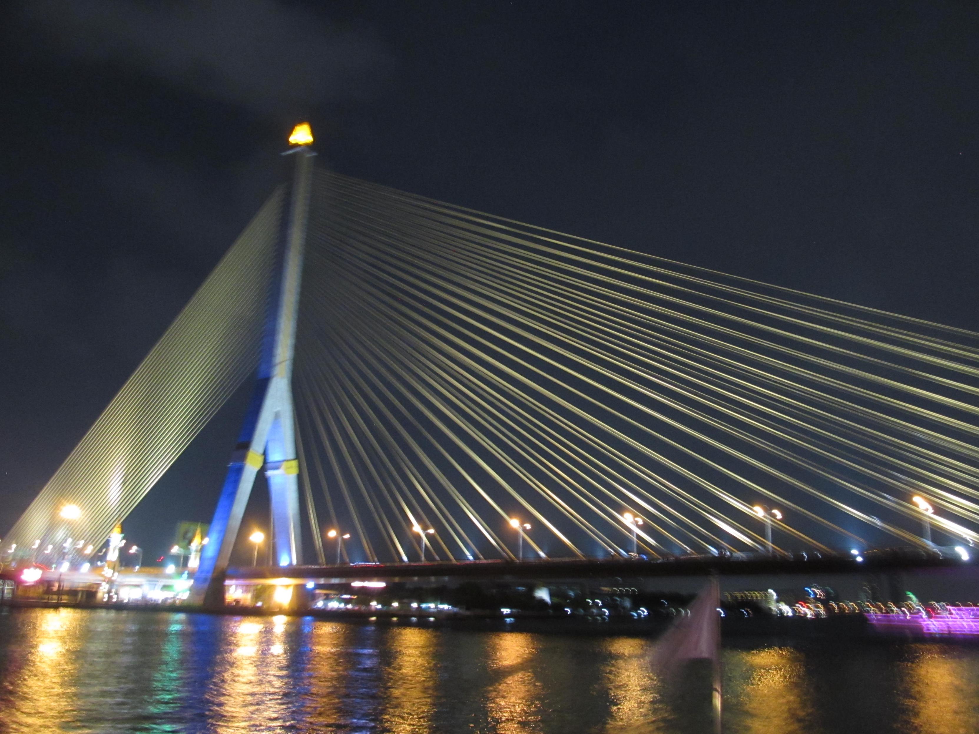 Nächtliche Aufnahme bei dunklem Himmel, im der Bildmitte schaut man auf eine hohe Schrägseilbrücke mit blauem Pylon, deren Seile von Scheinwerfern angestrahlt werden. im Vordergrund der Fluss, in dem sich verschiedene Farben widerspiegeln.