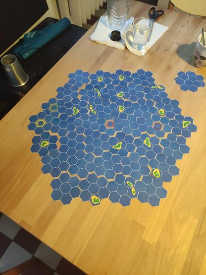 Auf einen Holztisch liegen Spielkarten mit einer sechseckigen Wabenstruktur mit viel Blau und ein paar kleineren "Inseln"