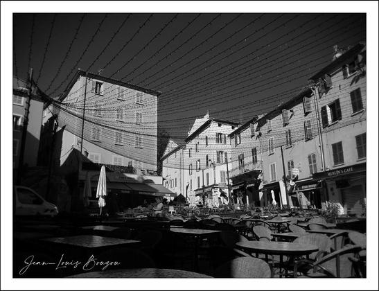 This black and white photograph beautifully captures a quiet, sunlit urban square framed by tall, classic European-style buildings. The architecture is characterized by simple, rectangular windows and gently sloping rooftops with chimneys, suggesting a historical or old-town ambiance. The shadows cast by the buildings and the uneven light filtering into the plaza evoke a serene, almost timeless atmosphere.
A striking visual detail is the array of thin, dark lines stretching diagonally across the image from building to building. These wires create a subtle grid or web above the plaza, adding a geometric rhythm and depth to the scene. This linear pattern juxtaposes the organic scattering of tables and chairs below, orderly but inviting, hinting at a community space—perhaps an outdoor café or market area.
The empty chairs and tables, all facing different directions, invite the viewer to imagine the lively conversations or quiet moments of solitude that might unfold here. Umbrellas stand folded and still, reinforcing the calm before the bustle of the day. A few parked cars nestle into the corners, emphasizing the blend of daily life and leisure.
Artistically, the monochrome treatment strips away distraction, focusing attention on shapes, contrast, and light play, while also invoking nostalgia or introspection. The signature “Jean-Louis Beque” at the bottom corners the image as a thoughtful artistic statement.
