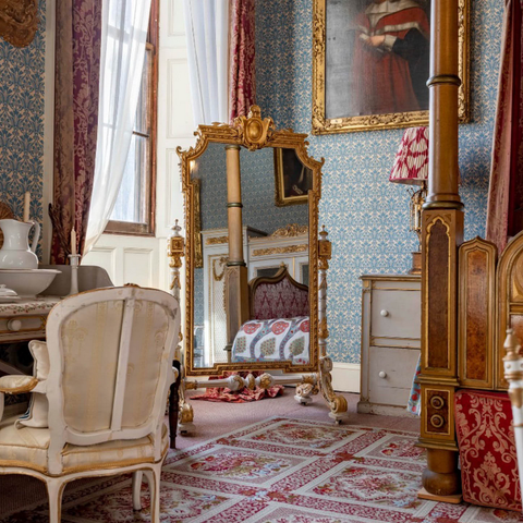 a photo of the corner of a bedroom, white drapes, a golden mirror, the post of the bed and a chair at the vanity, an intricate carpet underneath, white and red and pink and green 