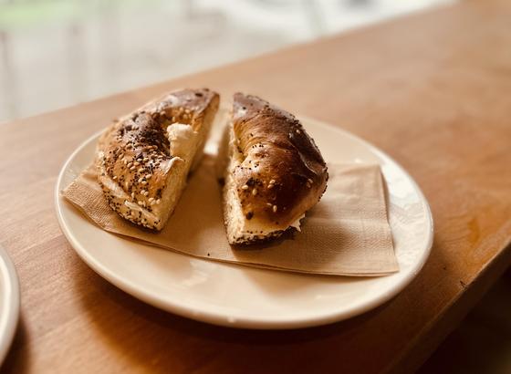 Ein getoasteter Bagel mit Sesam und Mohn bestreut. Er ist in zwei Hälften zerschnitten und mit Creamcheese gefüllt. Der Bagel liegt auf einer Serviette in einem Teller.