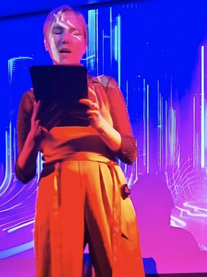 Aiki Mira mit Tablet liest aus Denial of Service zu glitchy Soundscapes von Michael Wehren vor der Projektion des neonpinken Buchcovers ♡