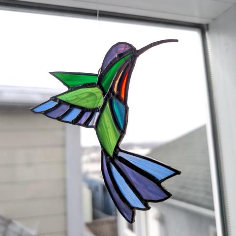 Photograph of a stained glass hummingbird that I designed and created, hanging in a window. The hummingbird design is based off of the fiery-throated hummingbird, with an orange throat, green and blue body, and purple-tinged wings. CC BY-SA Kate Zimmerman.