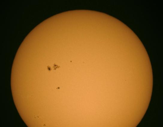 A close-up image of the Sun against a dark background, showcasing its bright orange surface with several dark sunspots visible.