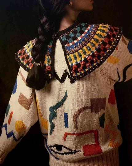Woman models a chunky cream knit sweater featuring colorful geometric patterns, a prominent studded Peter Pan collar, and an embroidered eye motif on the torso.