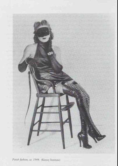 Black and white photograph of a woman wearing a black mask, halter dress, long gloves, and lace-up thigh-high stiletto boots, seated sideways on a wooden chair while holding a riding crop, circa 1944.