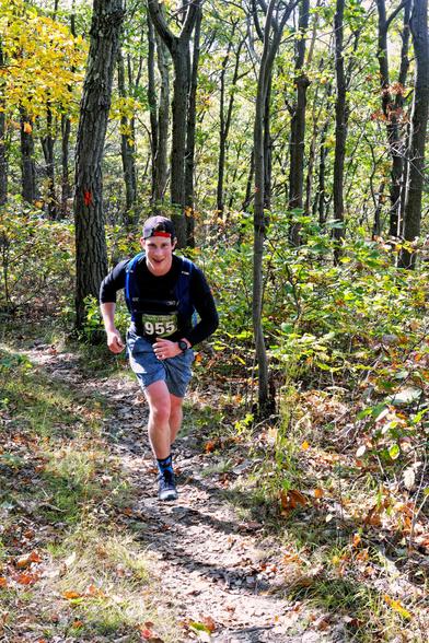A man with a headband is running up a sandy trail in a young forest with vigorous understory and just a few trees starting to turn color