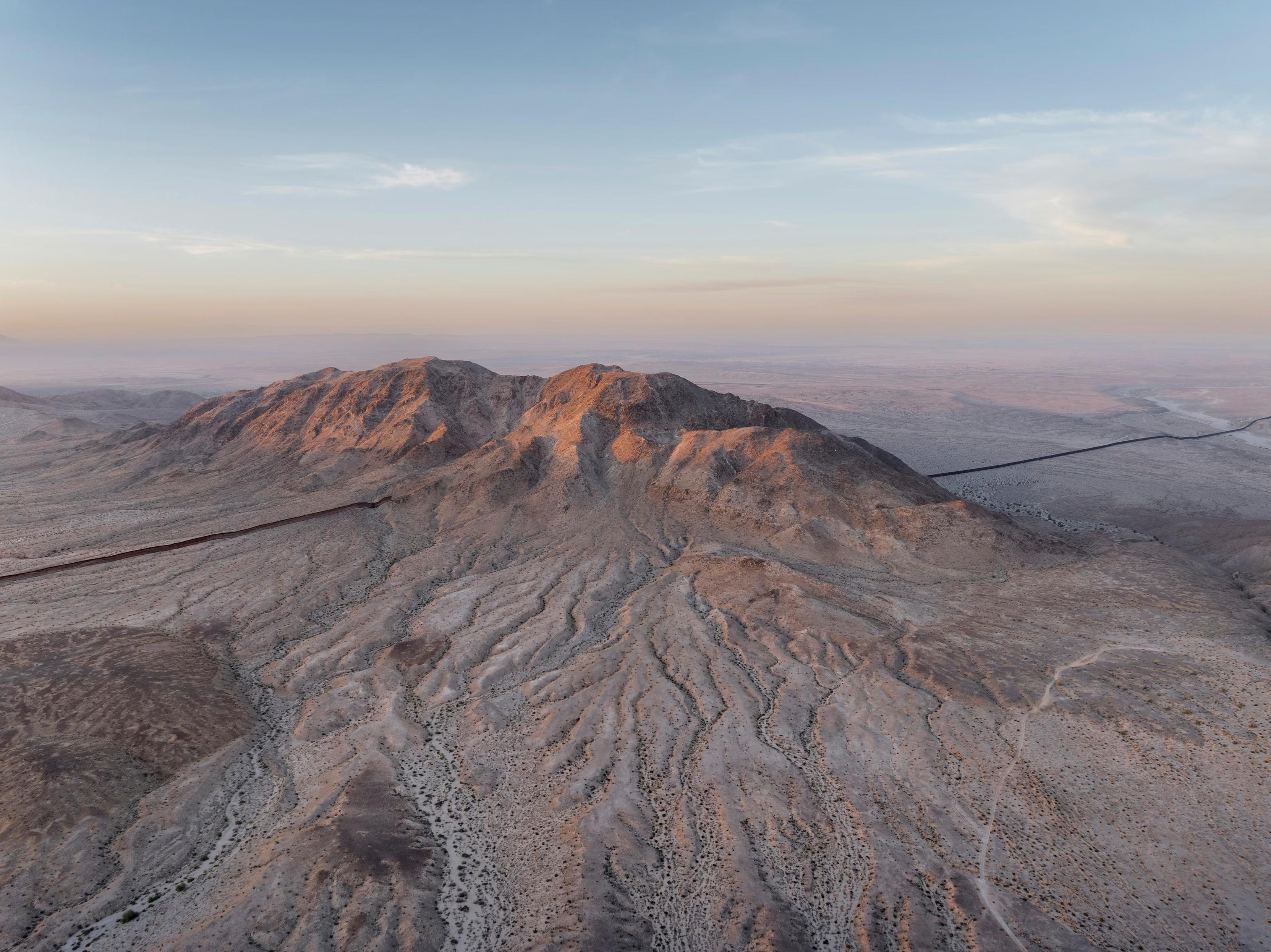 The setting sun illuminates a mountain that creates a gap in the border wall near Mexicali, Baja California