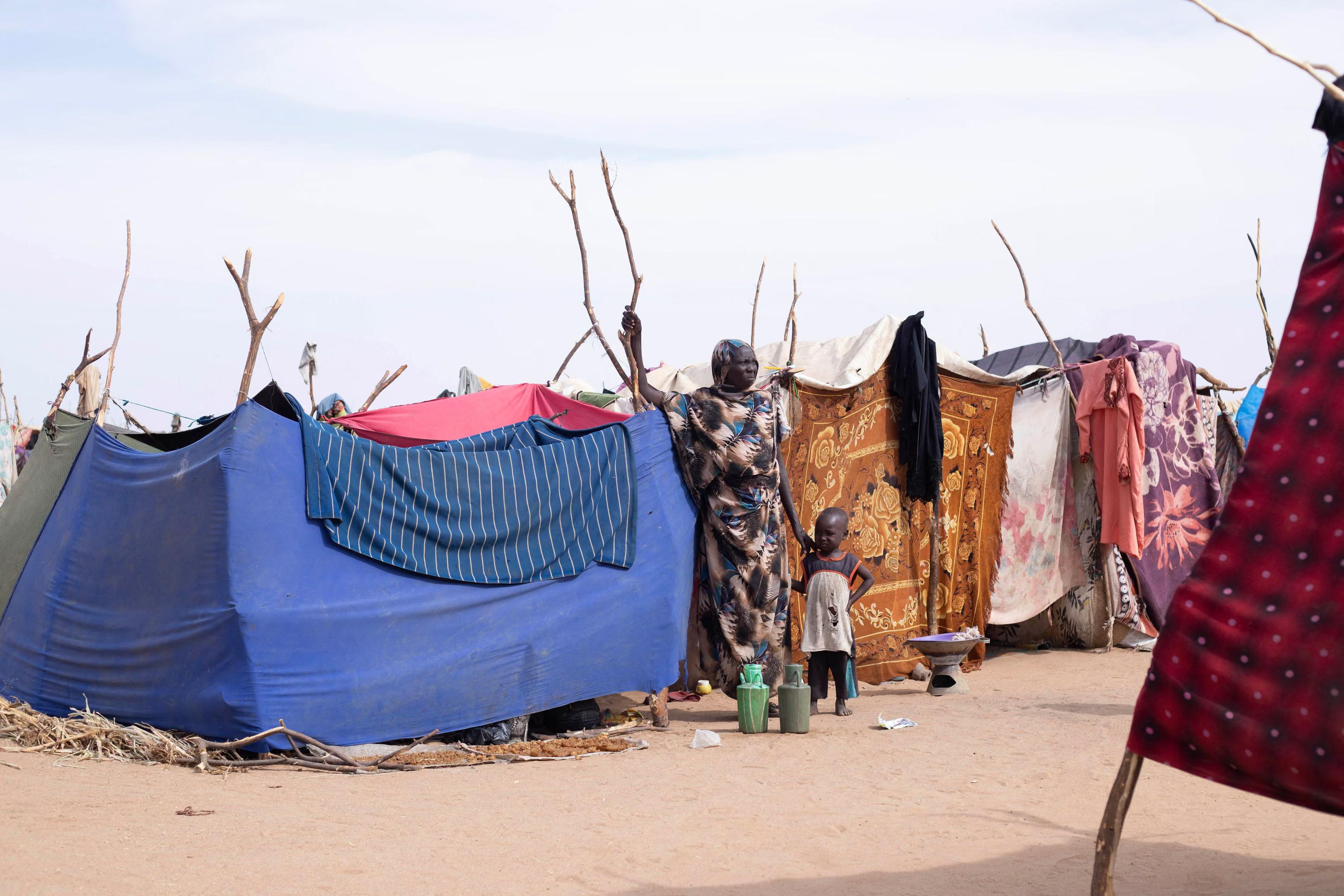 A displaced Sudanese woman and child stand in the Um Yanqur camp, in front of makeshift shelters.