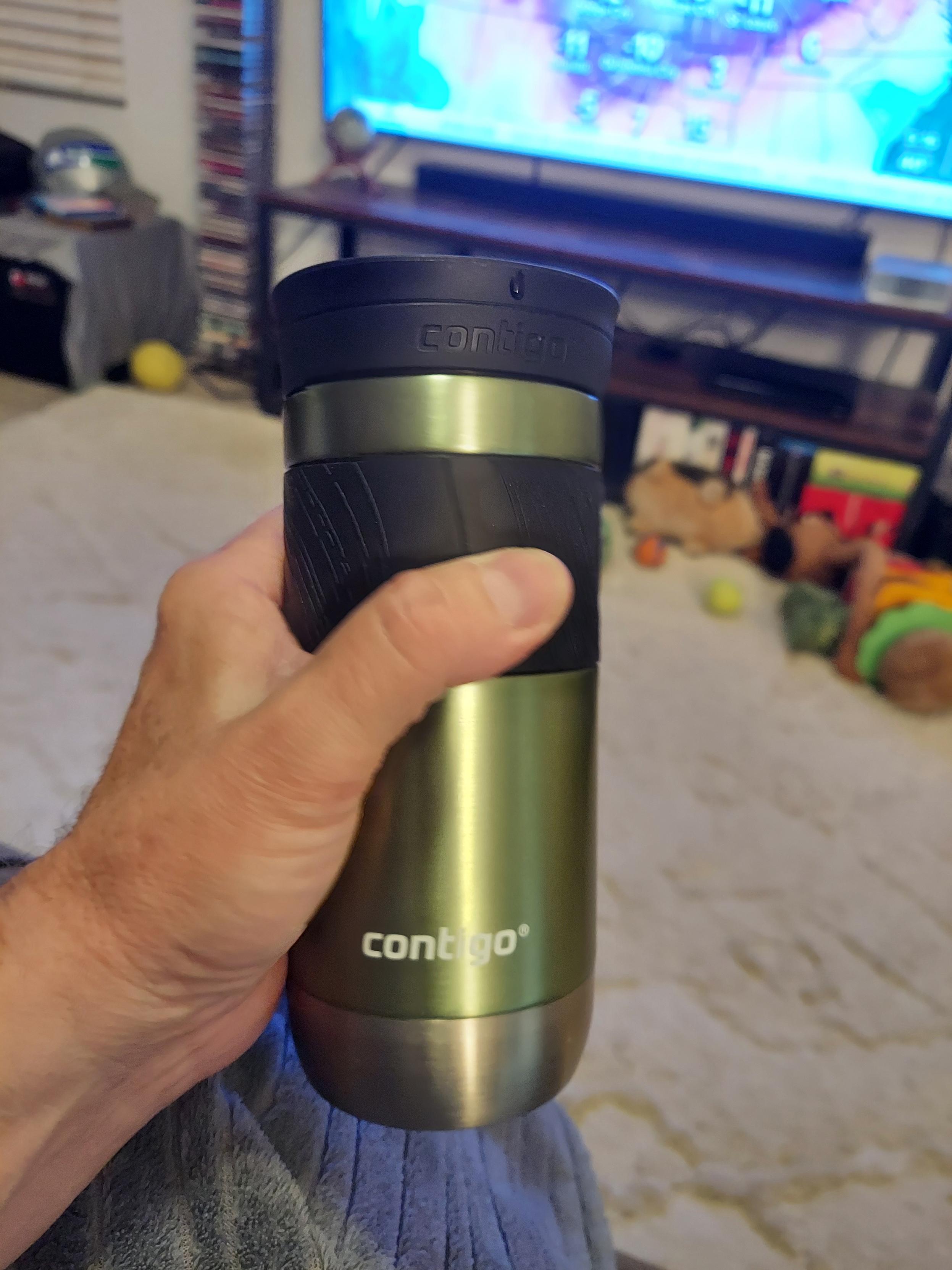 Photo of a left hand holding an insulated green and black aluminum beverage container. In the background is a running dumbbox, with scattered dog toys on the floor in front.
