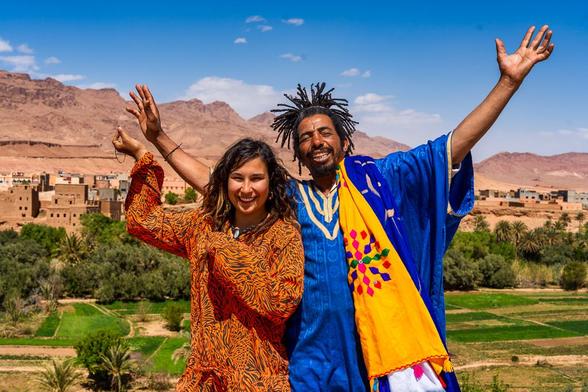 Eine lächelnde junge Frau und ein Mann mit Dreadlocks stehen im Sonnenschein vor einer beeindruckenden, trockenen Berglandschaft und einer traditionellen marokkanischen Siedlung.

Hinter dem Paar erstreckt sich eine üppige grüne Oase mit Palmen und bewirtschafteten Feldern, ein starker Kontrast zu den erdigen, roten und braunen Lehmbauten des kleinen Dorfes in der Mitte. Im Hintergrund dominieren die zerklüfteten, rötlichen Berge des Atlasgebirges die Szenerie unter einem strahlend blauen Himmel mit wenigen weißen Wolken. 