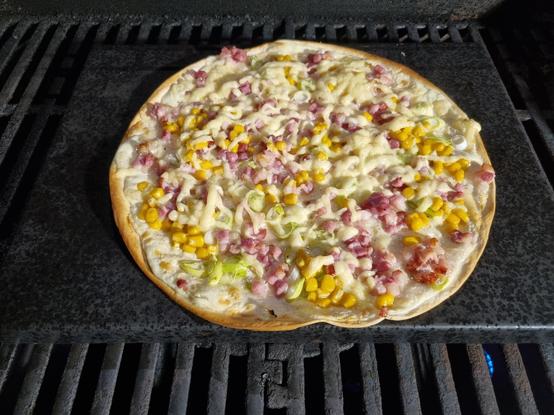 Ein Flammkuchen auf einem schwarzen Backstein auf einem Grillrost nähert sich dem perfekten Gargrad. Er ist mit Lauchzwiebeln, Speck und zur allgemeinen Erzürnung auch mit Mais belegt.