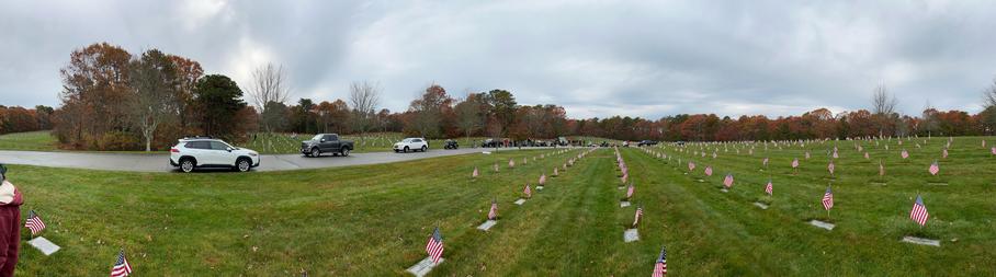 Panorama photo showing rolling hills on a rainy day, dotted with hundreds of small American flags placed over the headstones of US servicembers and their spouses buried 
A number of cars and a group of motorcycles can be seen parked on the circular drive that is in this section, belonging to the volunteers who showed up to place the flags