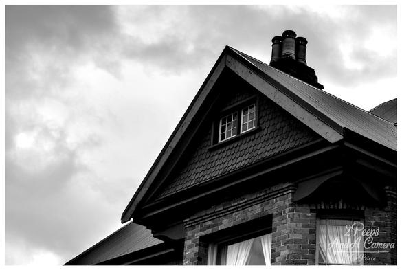 A dramatic black and white photograph, shot from a low angle, focusing on the steep gable and roof of a historic brick house in Queenstown.

The house features decorative scalloped shingles on the gable face, a small lattice window, and a prominent cluster of brick chimneys against a bright, cloudy, and moody sky.

The high contrast emphasises the texture of the brickwork and the metal roof.
