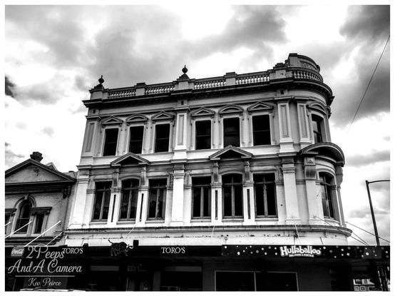 Black and white photograph of a stately, three story Victorian era building in Launceston, Tasmania, featuring arched windows and a prominent corner turret with a balustrade roofline against a cloudy sky.

The ground floor businesses include TORO'S and Hullaballoo.