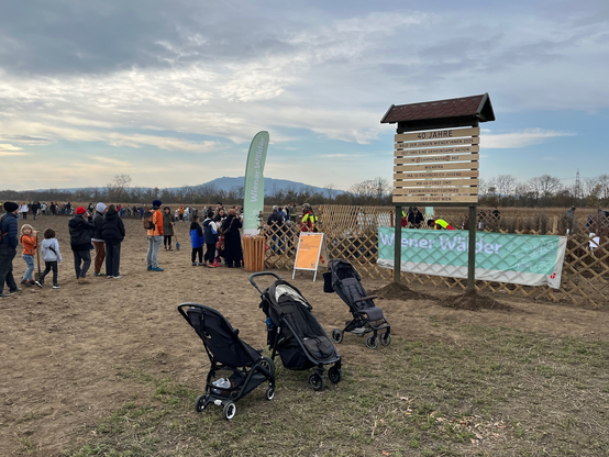 Ein Holzschild, das den Wald der jungen WIener*innen markert, davor abgestellt Kinderwägen, im Hintergrund Kinder & Erwachsene, die Bäume und Sträucher bei der Ausgabestelle nehmen um sie zu pflanzen