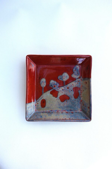 Quadratischer kleiner Teller mit dunkelroter und blaugrauer Glasur. Dargestellt ist ein Abhang mit 4 Bäumen.