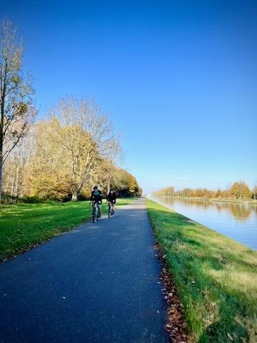 The fourth image features a straight, paved path running parallel to a long, calm body of water, likely a canal. Two cyclists, wearing dark clothing, are riding their bikes away from the viewer on the path. A strip of green grass separates the path from the water. On the left side of the path, there's more green grass and a line of trees with sparse, light-colored autumn foliage. The sky above is clear and blue, indicating a bright, sunny day.