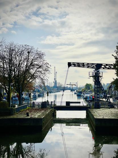 This image shows a serene canal scene with houses lining the left bank. The houses have various architectural styles, with some having white or dark facades and pitched roofs. Several boats, mostly small to medium-sized recreational vessels, are moored along the bank, reflecting in the calm, grey-green water of the canal. The sky above is overcast with soft, diffuse clouds, and a faint hint of trees with bare branches can be seen in the distance on the right.