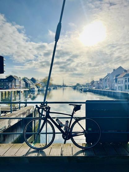 This image features a black road bicycle parked on a wooden bridge or platform, taking up most of the foreground. The bicycle is angled slightly, with its frame and wheels clearly visible. Beyond the bicycle, a canal stretches into the distance, flanked by rows of buildings on both sides. The architecture suggests a European town, with multi-story houses and other structures. The sky is partly cloudy, and the overall lighting suggests a clear day, with reflections shimmering on the water.