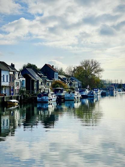 This photo presents a view looking down a canal towards the horizon, dominated by a bridge or lock structure in the mid-ground. The lock mechanism has large, dark metal components. On the left, a large tree with mostly bare branches stands. The canal water reflects the grey, cloudy sky. In the distance, numerous boats are visible along the banks, and further down the canal, there appear to be industrial structures and cranes silhouetted against the horizon, indicating a working waterway.
