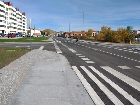 Foto de una intersección recién urbanizada. La acera finaliza súbitamente. Es necesario cruzar a la acera de enfrente para continuar caminando. Y tampoco hay acceso peatonal a la calle de la izquierda, donde se ven varios bloques de pisos.