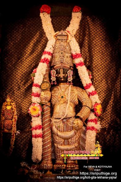 Closeup of idol of Hindu deity Krishna standing in diamond cladding with a matching crown, holding a similarly clad staff in right hand. A string of white flowers (some bands of red flowers) around Krishna floor-floor. Brown silk cloth with black cross-mesh pattern in the background.A string of white flowers hangs in U shape at the top of the background.