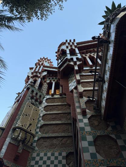 A detailed view of an architectural structure featuring red brick, colorful tiled patterns, and intricate design elements. The image captures the building's upper sections against a clear blue sky, framed by tree branches. The unique style suggests a blend of modernist and traditional styles. Casa Vicens in Barcelona designed by Gaudi.