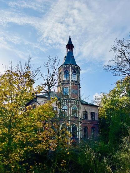 This image features a tall, ornate building, possibly a chateau or mansion, partially hidden by trees with autumn leaves. The building is made of brick and has a distinctive, multi-level tower topped with a steep, pointed spire. The tower has numerous windows with decorative framing, and intricate architectural details. The surrounding trees have a mix of green, yellow, and bare branches. The sky above is overcast with heavy grey clouds, creating a somber atmosphere.