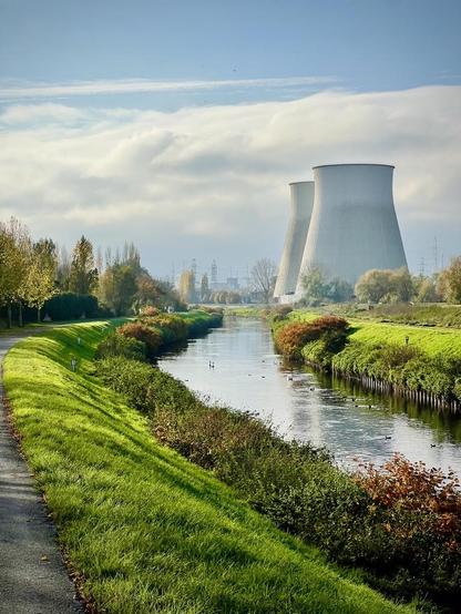 This image presents an industrial landscape with a natural foreground. A narrow canal or river flows from the foreground towards the distance, bordered by grassy banks on both sides. On the left bank, a paved path is visible, alongside trees with some autumn foliage. In the mid-ground, dominating the right side of the image, are two large, grey, conical cooling towers, characteristic of a power plant or industrial facility, partially obscured by a hazy, overcast sky. Further industrial structures can be faintly seen in the distance, blended into the foggy backdrop.
