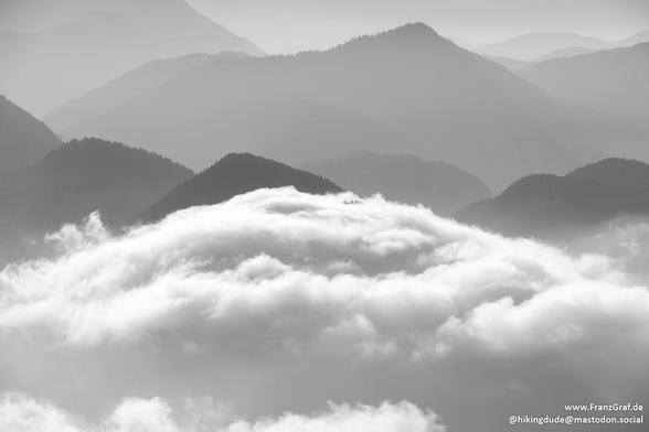 A sea of soft, billowing clouds stretches out beneath a dramatic mountain landscape, their fluffy white peaks gently undulating like waves. The mountains rise majestically from the cloud cover, their rugged silhouettes softened by the misty atmosphere. Layer upon layer of forested ridges and peaks fade into the distance, creating a sense of depth and scale that feels almost infinite.

The scene is serene and ethereal, with the clouds obscuring the lower slopes and adding an air of mystery to th…