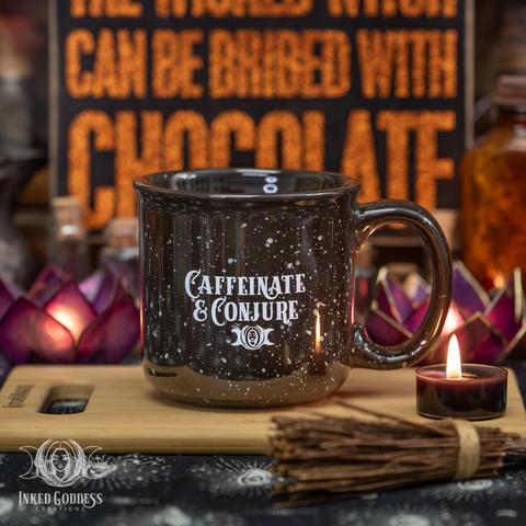 One "Caffeinate & Conjure Ceramic Mug" sits on a small bamboo cutting board. It's glazed black with white specks throughout. The lip is slightly flared. It has "Caffeinate & Conjure" printed on the side along with a small IGC logo below. This is an exclusive product from Inked Goddess Creations.