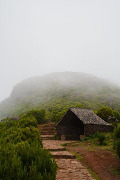Auf einem Gipfel steht in Nebel gehüllt ein kleines Haus, zu dem ein rötlicher Weg hinführt.