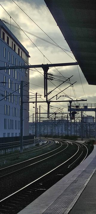 Voie ferrée urbaine au crépuscule, les rails formant une courbe élégante sous un ciel nuageux. Les câbles et structures métalliques se découpent en ombres fines sur la lumière diffuse, créant une atmosphère calme et mélancolique de gare en suspension.
