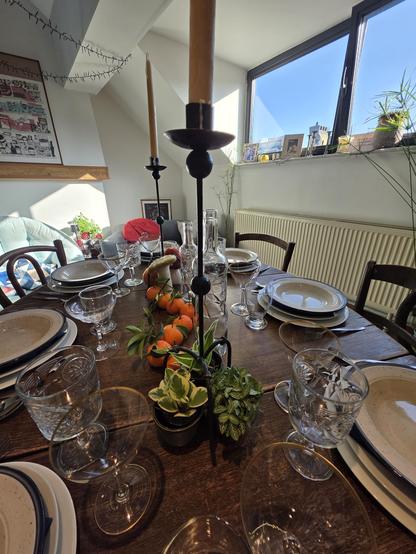 Voici une vue en plongée d’une table en bois foncé dressée pour un repas. Au centre de la table se trouvent deux chandeliers noirs avec des bougiee jaunes. En dessous et entre les chandeliers, il y a un trio de petites plantes vertes, des mandarines placées en zigzag et de faux champignons. La table est dressée pour plusieurs convives avec des assiettes blanches et des verres à vin sur chaque place. Le fond de l'image montre une fenêtre avec vue sur un paysage urbain et un ciel bleu.
