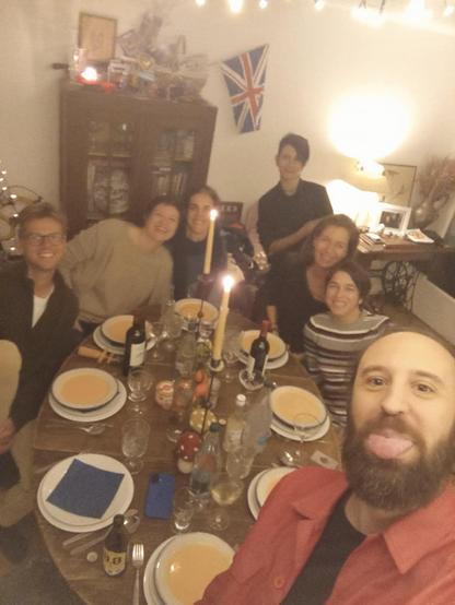 Une personne au premier plan regarde directement la caméra et tire la langue en  souriant. Six autres personnes sont assises autour d'une table en bois, et des plats sont devant chacune d'entre elles. Sur la table se trouvent des bouteilles, des verres et des bougies allumées. Derrière les personnes, un meuble brun foncé est rempli d'objets divers. L'éclairage est sombre, mis à part la lueur des bougies.