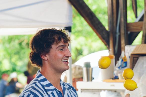 Young man with short hair wearing a blue and white thick striped shirt is seen shoulders-up and smiling,  the are three lemons hovering in the air in front of him the scene behind is a little confused but is mostly green leaves of trees,  tourists, wood framework and some kind of fabric sign or screen