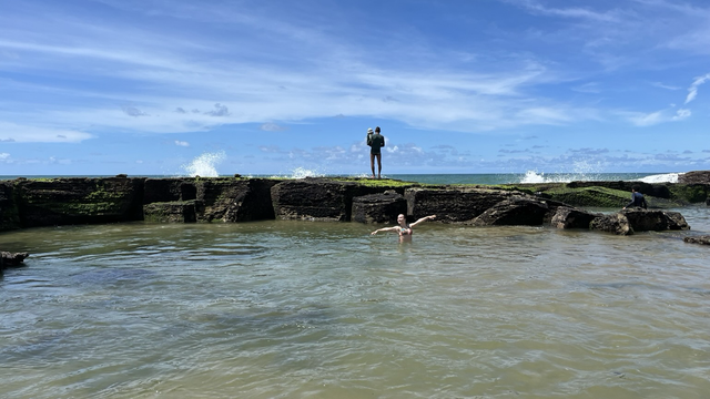 Un océano de mar transparente, es como una piscina entre rocas , se ve una joven en el mar y detrás, encima de las rocas, una persona