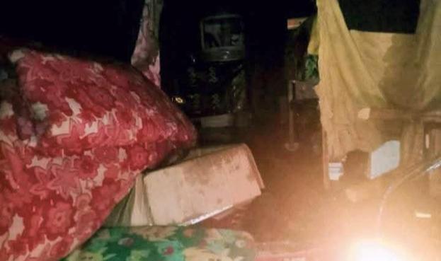 The image depicts a dark, dimly lit indoor that appears to be flooded or severely messy. On the left, a large piece of tan or yellow fabric hangs. In the center, a dark large furniture item, is partially visible in the shadows. The floor is covered in dark water or mud. On the right, a large object covered in a reddish fabric with a white floral pattern dominates the foreground, suggesting a mattress or large cushion.