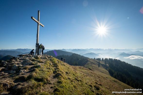 A majestic summit unfolds beneath a brilliant blue sky, where the sun shines brightly, casting a radiant glow over the landscape. At the peak, a wooden cross stands tall, serving as a focal point and symbol of achievement for those who reach this breathtaking viewpoint. The cross is surrounded by a small group of hikers, their figures silhouetted against the stunning backdrop of rolling hills and distant mountain ranges.

The panoramic view stretches out in layers of forested ridges and valleys…