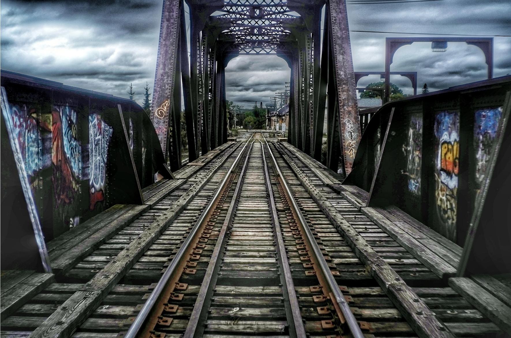 Walking along the center of train tracks crossing a bridge. Graffiti can be seen on either side of the built up sides of the metal bridge as well as the tall steel structure further along. Moody clouds fill the sky.