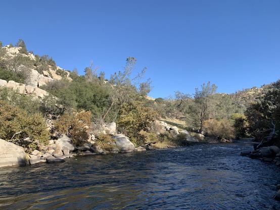 A River, seen from its bank, flows between rocky hills with trees on a sunny day