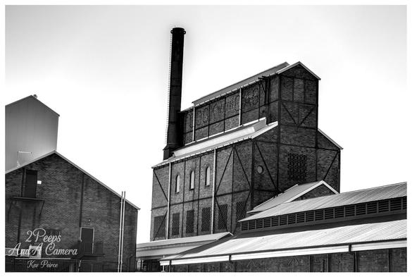 A high contrast black and white photograph of the historic Launceston Gasworks buildings. The central structure is a large, dark brick industrial building with external steel framing and a tall, slender chimney rising from the top. 

Smaller brick and corrugated iron buildings are visible on either side and in the foreground. The bright white sky provides a dramatic background.