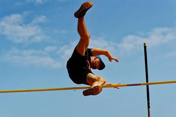 A man is frozen in air with hisfeet facing us and his hands still behind the yellow horizontal bar,  a green pole that got him there is resting on the far side and there are thin clouds in the far distance